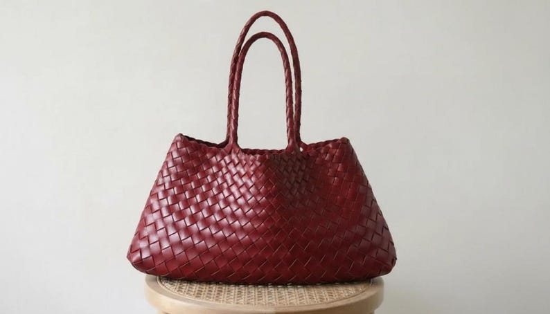 May include: A burgundy tote bag with woven detailing. The bag has two braided handles and sits on a small, round stool. The bag's woven pattern is visible across its body. The background is a plain, off-white wall.