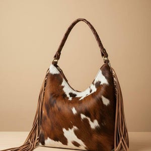 May include: A brown and white cowhide shoulder bag with a braided brown leather handle. The bag features long brown fringe on the sides. The bag is sitting on a light brown surface.