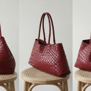 May include: A set of three burgundy-colored tote bags. Each bag features a woven design and two handles. The bags are displayed on small, round wooden stools with woven seats. The background is a plain, light-colored wall.