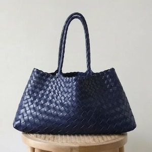 May include: A navy blue woven tote bag with two handles. The bag is made of a woven material and sits on a small wooden stool. The bag has a trapezoid shape and is empty.