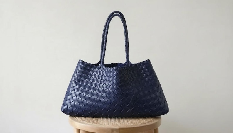 May include: A navy blue woven tote bag with two handles. The bag is made of a woven material and sits on a small wooden stool. The bag has a trapezoid shape and is empty.
