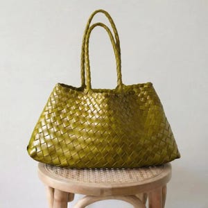 May include: A woven olive green tote bag with braided handles. The bag is made of a woven material and has a trapezoid shape. The bag is sitting on a small, round, light-colored wooden stool.