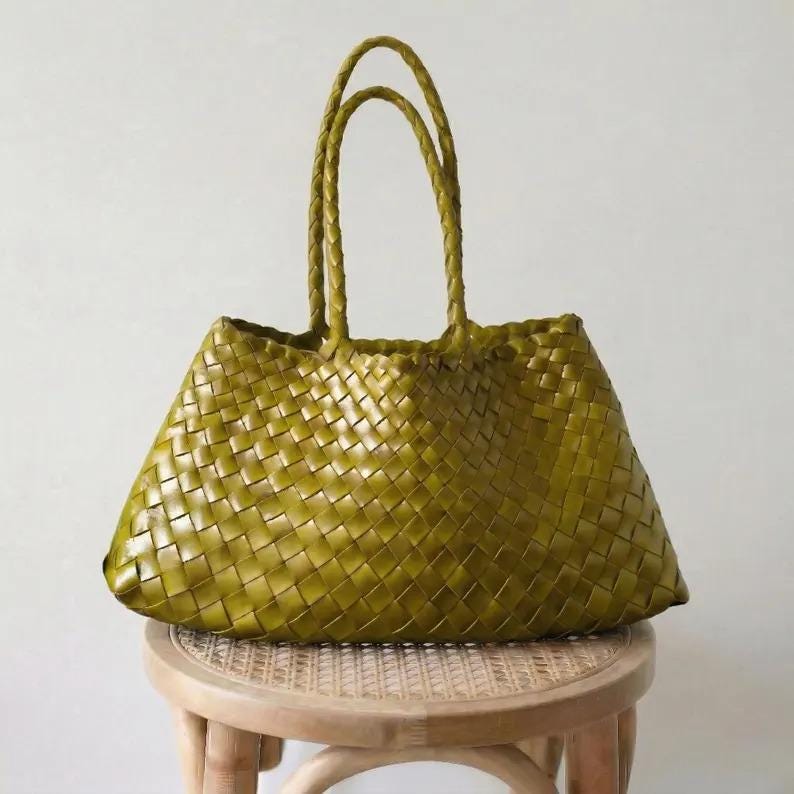 May include: A woven olive green tote bag with braided handles. The bag is made of a woven material and has a trapezoid shape. The bag is sitting on a small, round, light-colored wooden stool.