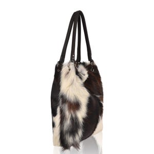 Puede incluir: Bolso tote de piel de vaca con una mezcla de pelo negro, marr&oacute;n y blanco. El bolso tiene dos asas de cuero marr&oacute;n oscuro y anillos met&aacute;licos plateados. Dise&ntilde;ado para llevar al hombro.