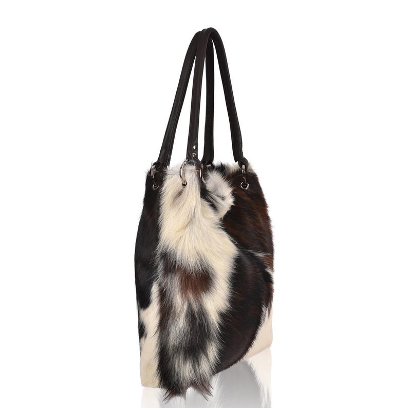 Puede incluir: Bolso tote de piel de vaca con una mezcla de pelo negro, marr&oacute;n y blanco. El bolso tiene dos asas de cuero marr&oacute;n oscuro y anillos met&aacute;licos plateados. Dise&ntilde;ado para llevar al hombro.