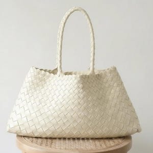 May include: Cream-colored woven tote bag with two braided handles. The bag has a trapezoid shape and is sitting on a small, round, wooden stool. The weave pattern is visible across the entire surface of the bag.