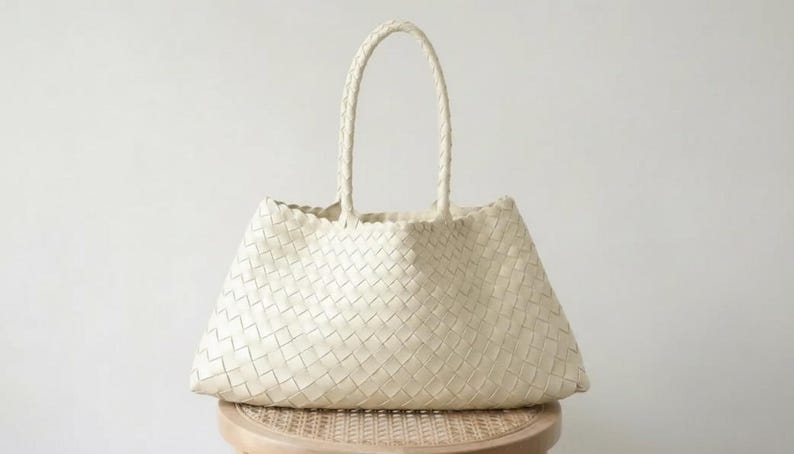 May include: Cream-colored woven tote bag with two braided handles. The bag has a trapezoid shape and is sitting on a small, round, wooden stool. The weave pattern is visible across the entire surface of the bag.