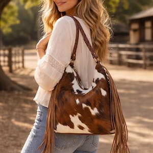 May include: A brown and white cowhide shoulder bag with long brown fringe. The bag has a braided brown leather strap and gold-colored hardware. The woman is wearing a white blouse and blue jeans.