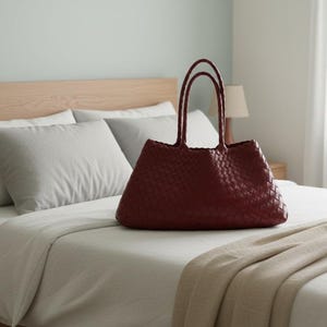 May include: A burgundy woven tote bag with double handles sits on a bed with white linens and gray pillows. The bag has a structured shape and a textured surface. The room has a light, airy feel with a wooden headboard and a neutral color palette.