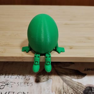 Può includere: Una figura verde brillante a forma di uovo, stampata in 3D, con piccole membra. La figura è seduta con la testa china e le gambe penzolanti. L'oggetto è in plastica e ha una superficie strutturata.