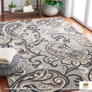 May include: A large, rectangular rug with a detailed paisley pattern in shades of grey, beige, and dark brown. The rug features a raised, textured design and is placed on a wooden floor. The rug is a decorative home decor item.