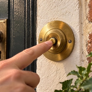 Peut inclure: Une sonnette de porte circulaire en laiton avec un bouton-poussoir central, fixée sur un mur blanc texturé. La sonnette présente une finition métallique et est fixée par deux vis visibles. Elle est située près d'une porte sombre avec une poignée.