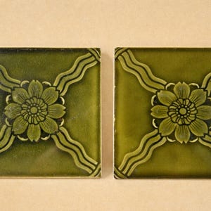 Puede incluir: Dos baldosas cuadradas de color verde oliva con un diseño floral. Cada baldosa presenta una flor central con pétalos detallados y un patrón circundante de líneas curvas. Las baldosas son probablemente para uso decorativo.