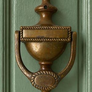 May include: A brass door knocker with a weathered patina, featuring a central urn-shaped body, a rectangular top plate with rope-style trim, and a circular bottom plate with matching rope detailing, mounted on a sage green paneled door surface.