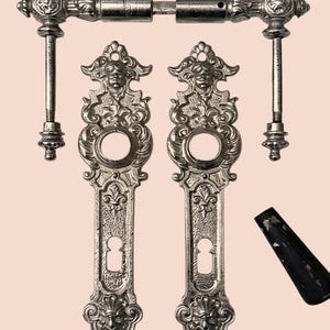 Könnte beinhalten: Antikes Türbeschlagsset in Silberoptik. Enthält zwei verzierte Türgriffplatten mit Schlüssellöchern, eine Verbindungsstange und einen separaten schwarzen Griff. Die Platten weisen detaillierte Gesichts- und Blumendesigns auf.