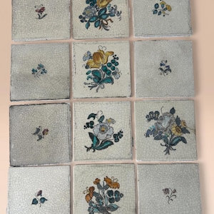 May include: Twelve square ceramic tiles with floral patterns. Each tile displays a unique hand-painted flower arrangement in yellow, blue, orange, and white on a cracked, off-white surface. The tiles are arranged in a grid formation.