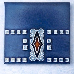 Può includere: Piastrella quadrata in ceramica blu con un disegno decorativo centrale. Il disegno presenta una forma a diamante in marrone e bianco, circondata da motivi a spirale. Piccoli accenti quadrati bordano il disegno centrale e i bordi.