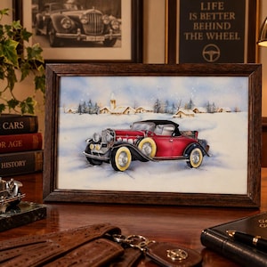 Könnte beinhalten: Ein gerahmtes Aquarellbild, das ein rotes Oldtimer-Cabriolet mit schwarzen Kotflügeln und Weißwandreifen zeigt, das in einer verschneiten Landschaft mit kleinen Gebäuden im Hintergrund unter einem hellblauen Himmel geparkt ist.