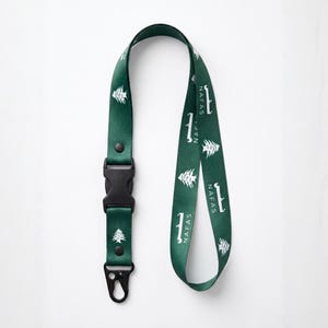 May include: A dark green lanyard featuring a repeating white graphic of a cedar tree and the text NAFAS in both English and Arabic script. The lanyard includes a black plastic side-release buckle and a black metal lobster claw clasp attachment.