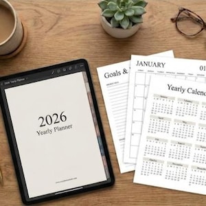 Op de afbeelding: Een zwarte tablet met een jaarplanner voor 2026, samen met afgedrukte jaarlijkse kalenderpagina's en een gouden pen. Een kopje koffie, een kleine vetplant en een bril liggen op een houten oppervlak.