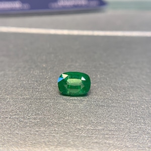 May include: A faceted, oval-shaped emerald gemstone. The gem is a vibrant green color and sits on a gray surface. The cut of the emerald creates multiple reflective surfaces, enhancing its brilliance. The gemstone is a focal point.