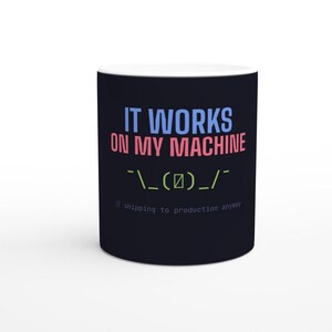 Könnte beinhalten: Ein schwarzer Keramikbecher mit dem hellblauen Text IT WORKS, dem rosafarbenen Text ON MY MACHINE, einem grünen Achselzucken-Emoticon und dem grauen Text // shipping to production anyway. Das Design ist mittig auf der zylindrischen Tasse platziert.