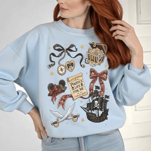 Könnte beinhalten: Hellblaues Sweatshirt mit Piratenmotiven. Das Design umfasst eine Schatzkiste, ein Piratenschiff, gekreuzte Schwerter und eine Schriftrolle mit dem Text "A Pirate's Life For Me". Akzente mit Schleifen, Sternen und Mickey Mouse-Silhouetten.