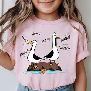 May include: Pink t-shirt featuring a cartoon design of two seagulls on a rock, with the word "MOM!" repeated around them. A small orange fish is in the water below. The shirt is made of a soft material.
