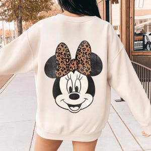 May include: Cream-colored sweatshirt featuring a large Minnie Mouse graphic on the back. The graphic includes a black and white Minnie Mouse face with a leopard print bow. The sweatshirt is a casual, comfortable style.