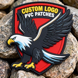 May include: A PVC patch featuring a detailed eagle design with outstretched wings. The eagle is black, white, and blue, set against a red shield-shaped background. The text "CUSTOM LOGO PVC PATCHES" is in yellow. The patch is on a rocky surface.