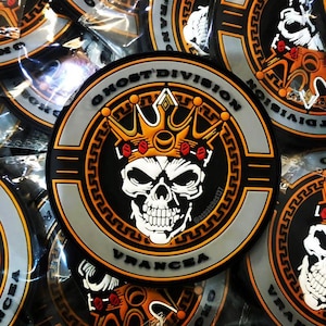 Puede incluir: Parche circular con una calavera coronada, con el texto "GHOST DIVISION VRANCEA" que la rodea. El diseño incorpora elementos en negro, naranja y gris. La calavera es blanca.