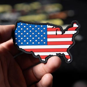 Peut inclure: Un écusson en forme de carte des États-Unis, représentant le drapeau américain. Le design comprend des rayures rouges et blanches et une section bleue avec des étoiles blanches. L'écusson a une bordure noire.