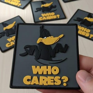 May include: Square patch featuring Daffy Duck with the text "WHO CARES?" in yellow. The patch has a black border and a gray background. Several similar patches are visible in the background.
