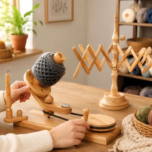 Può includere: Un avvolgitore per lana in legno chiaro con una matassa di filato grigio scuro, dotato di manovella manuale e meccanismo a cinghia. Accanto si trova un arcolaio in legno, uno strumento regolabile per tenere in posizione le matasse di filato.