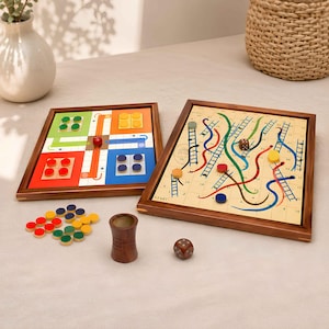 May include: A wooden board game set featuring a square Ludo board with red, green, yellow, and blue sections, and a rectangular Snakes and Ladders board with a numbered grid, colorful paths, and ladders. Includes wooden tokens, a dice cup, and dice.