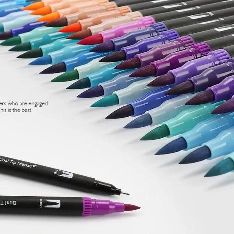 Op de afbeelding: Een verzameling dual tip markers gerangschikt in een diagonale rij, met zwarte behuizingen en witte tekst Dual Tip Marker. Elke marker heeft een gekleurde penseelpunt aan het ene uiteinde en een fijne punt aan het andere uiteinde.