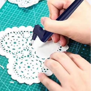 Puede incluir: Un cúter azul oscuro corta un tapete de papel blanco sobre una esterilla de corte verde. El tapete tiene un delicado diseño de encaje. El cúter se sostiene en una mano, demostrando su uso para manualidades en papel.