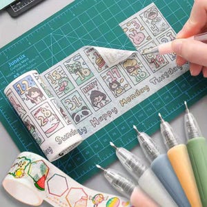 May include: A roll of decorative washi tape featuring a grid of numbered calendar dates, cartoon characters, and the text "Sunday Happy Monday Tuesday". A second roll shows hexagonal frames and food illustrations. Five gel pens are arranged in a row.
