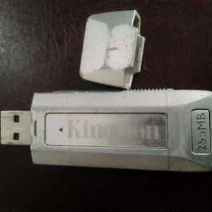 Puede incluir: Una memoria USB Kingston plateada con una etiqueta de capacidad de almacenamiento de 256MB y una tapa extraíble. El dispositivo rectangular muestra signos de desgaste en su superficie metálica y tiene un conector USB estándar y una anilla.
