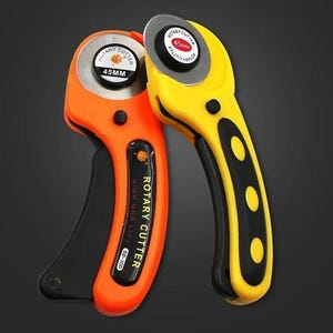 Puede incluir: Dos cortadores rotativos con cuchillas circulares de 45 mm. Uno tiene un mango naranja con detalles negros y el texto "ROTARY CUTTER SD-100", mientras que el otro tiene un mango amarillo con detalles negros. Ambos incluyen un mecanismo de seguridad.