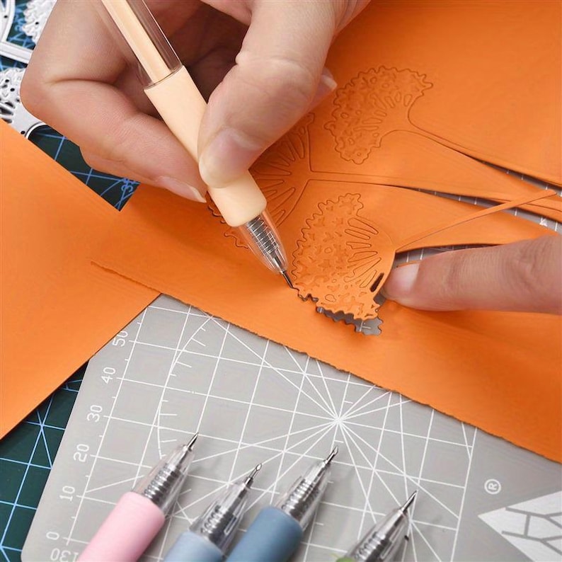 May include: A light peach-colored craft knife with a fine metal tip is being used to carefully cut out a detailed orange paper cutout featuring a dandelion design. Several other craft knives in pastel colors are arranged on a gray cutting mat.