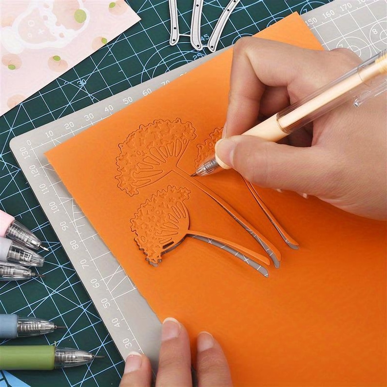 May include: A light-colored mechanical pencil held over a sheet of orange paper featuring intricate, laser-cut floral designs. The paper rests on a green cutting mat with white grid lines, alongside several other pens and metal craft dies.