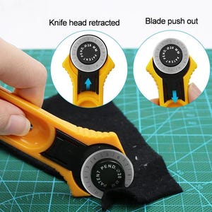Può includere: Un taglierina rotativa gialla con una lama circolare da 28 mm che taglia tessuto nero. Due riquadri circolari mostrano il meccanismo: uno etichettato Knife head retracted con una freccia verso l'alto e uno Blade push out con una freccia verso il basso.