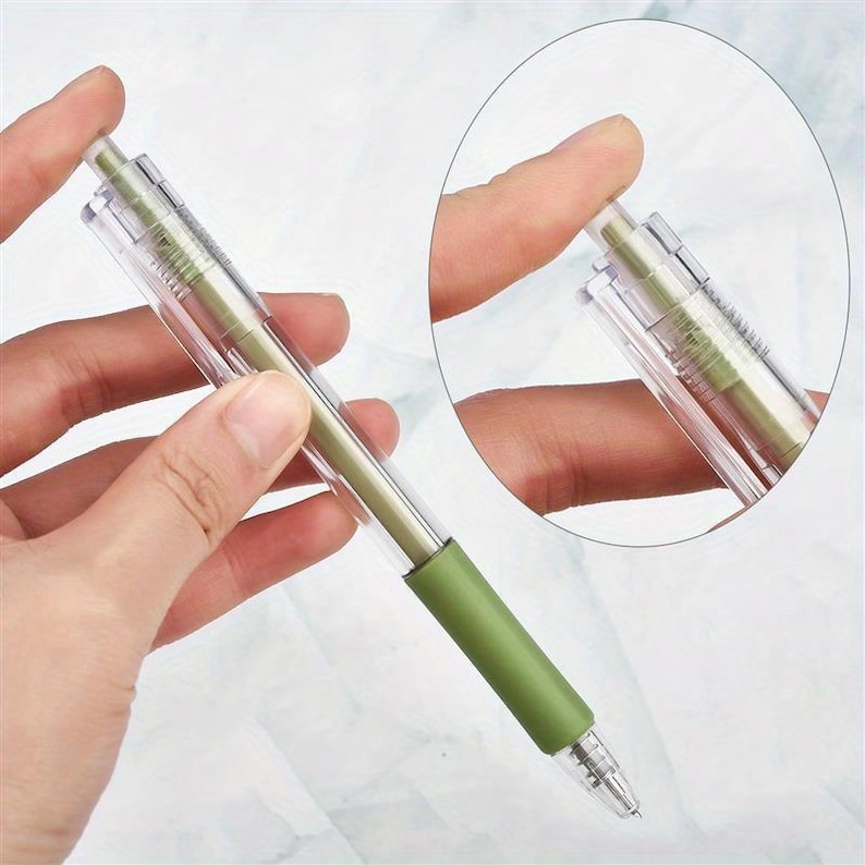 May include: A retractable ballpoint pen featuring a clear plastic barrel, a sage green internal ink cartridge, and a matching sage green rubberized grip section near the tip. The pen includes a clear pocket clip and a push-button mechanism at the top.