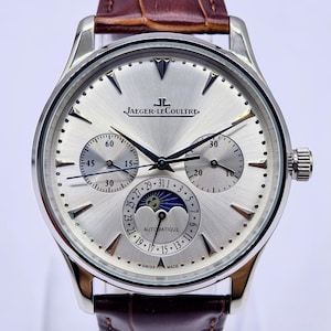 May include: Silver Jaeger-LeCoultre watch with a brown leather strap. The watch face has multiple sub-dials and a moon phase indicator. The watch is marked "AUTOMATIQUE" and "SWISS MADE".