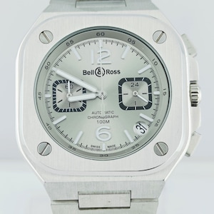 May include: A silver Bell & Ross automatic chronograph watch with a square face. The watch has a silver dial with black numerals, two sub-dials, and a date display. The watchband is made of silver metal links.