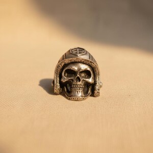 May include: A silver-colored ring featuring a detailed skull wearing a helmet. The helmet has a crest on the front. The ring is set against a neutral background, highlighting the intricate design and metallic sheen of the jewelry.