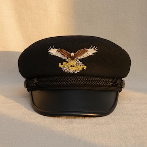 Puede incluir: Gorra negra con visera de cuero y banda trenzada. La gorra presenta un emblema de águila bordado con las palabras "Liberty Justice" en dorado. La gorra tiene una corona redondeada y un diseño estructurado.