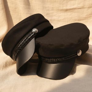 May include: Two black sailor hats with a braided band and a silver button detail. The hats have a structured shape with a curved brim. The hats are made of a soft fabric and have a leather-like brim.