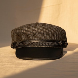 Peut inclure: Casquette de marin grise et noire à motif pied-de-poule avec une visière noire. Le chapeau est orné d'une bande décorative et de deux boutons argentés. Un accessoire de mode élégant.